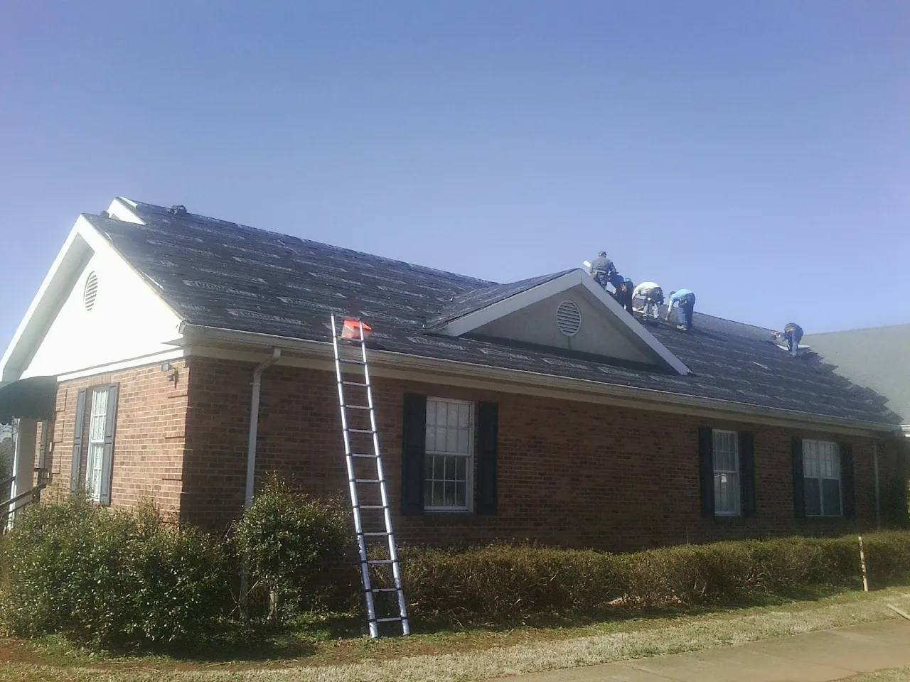 Crew tearing shingles off a brick ranch roof with a ladder set — Emergency Roof Repair work by our crew in Lodge