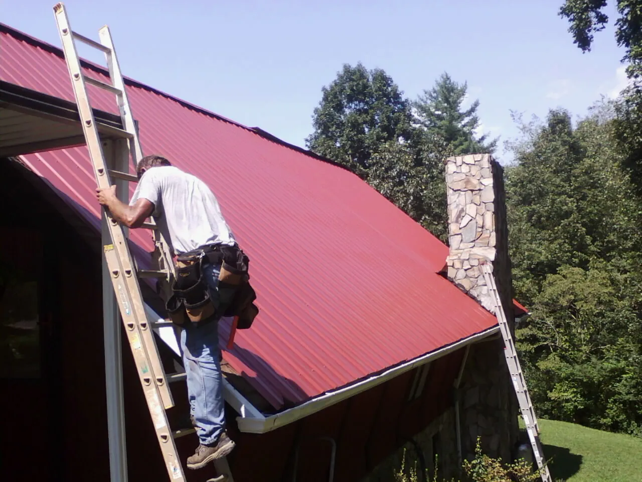 Roofer on a ladder beside a red metal roof and stone chimney — Soffit & Fascia Repair by Roofing Brothers in Lodge